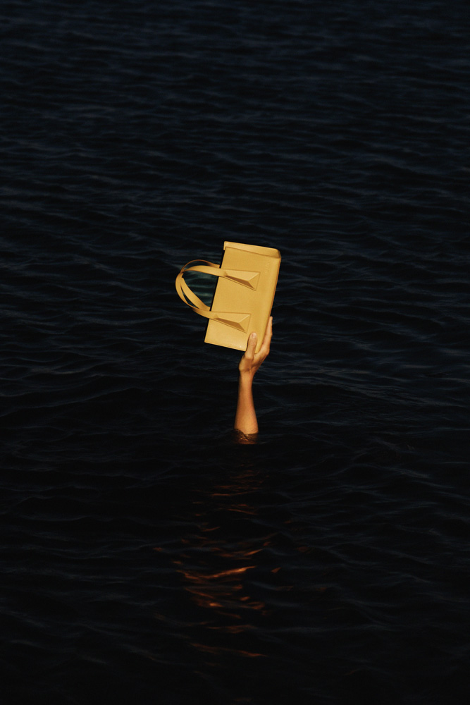 A tan purse is held aloft by an arm emerging from dark water, creating a striking contrast of light and dark.