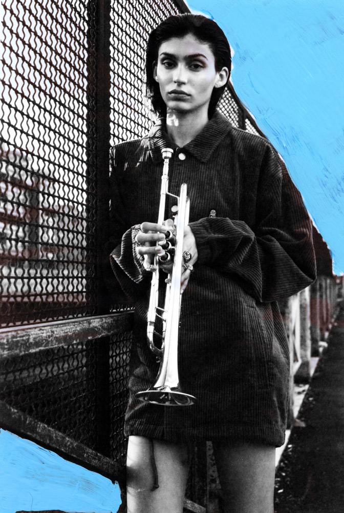 A young adult woman in a black corduroy jacket holds a trumpet, standing against a chainlink fence in a stylized urban scene.
