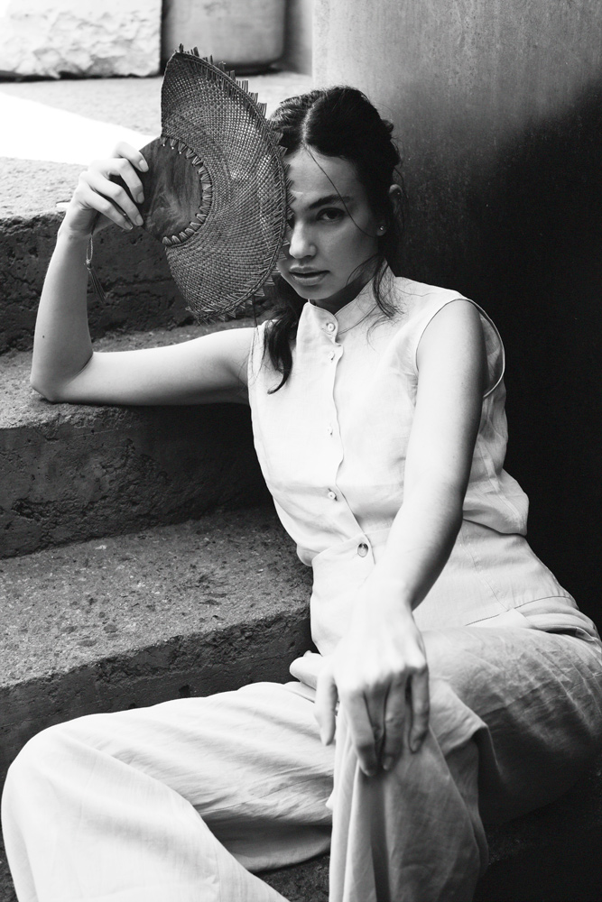 Woman in sleeveless top and pants sits on concrete steps holding a woven hat, in a black and white fashion-style photo.