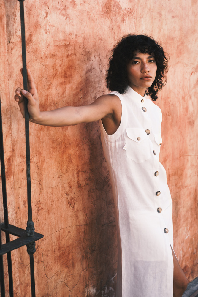 A woman poses in a sleeveless white button-down dress, holding onto a black metal railing against an orange stucco wall with shadows.
