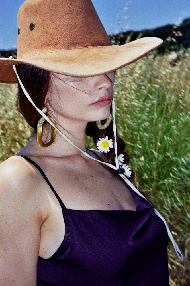 Woman in cowboy hat and earrings poses in a field. Braided hair is adorned with white flowers. Close-up shot with natural lighting.