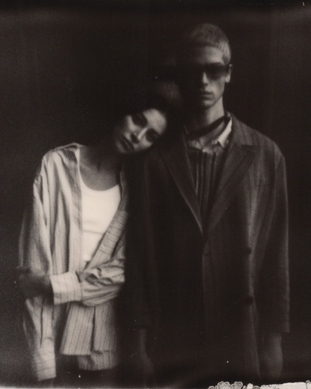 Monochrome fashion portrait of an adult male and female couple, wearing striped shirts, a jacket, and sunglasses, in a photo studio.