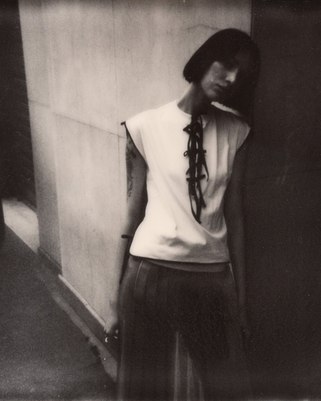 Monochrome portrait of a young woman in a white top with black laces, standing against a wall, with short dark hair and subtle expression.