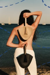 Woman with long dark hair holding two fan-shaped leather handbags by the ocean wearing white textured trousers
