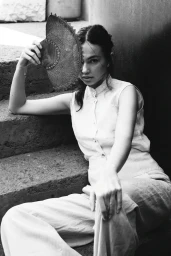 Monochrome portrait of a woman sitting on stone stairs holding a round woven fan in front of her face.