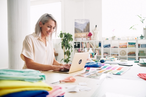Smiling woman with short blonde hair is using a laptop at a table covered in fabric, color swatches and sewing tools in an office.