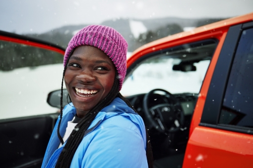 A smiling Black woman in a pink beanie and blue jacket is standing by an orange SUV in a snowy, mountainous landscape, eye-level shot.