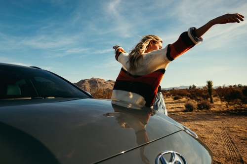 Blonde woman leans out of a car hood with arms spread wide in the desert under a blue sky with scattered clouds and rocky hills.