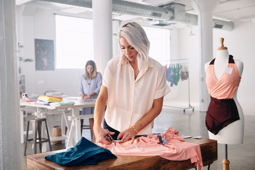 Fashion designer working at wooden table in studio, cutting pink fabric with scissors next to a dress form with garment.