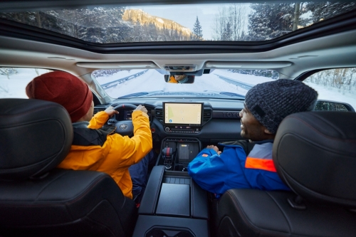 Inside a car driving on a snowy road: two people, one wearing an orange jacket, another in blue, seen through the open sunroof and windshield.