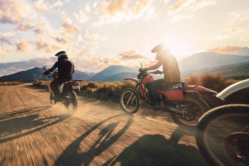 Three motorcyclists ride on a dirt road through a dry landscape with mountains in the background and a cloudy sky at sunset.