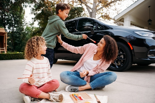 Mother and two children playfully paint near a black car in a driveway. The family is outdoors on a sunny day, smiling.
