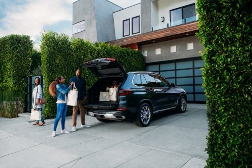 Family loads shopping bags into dark SUV parked outside modern house with green hedges and garage door.