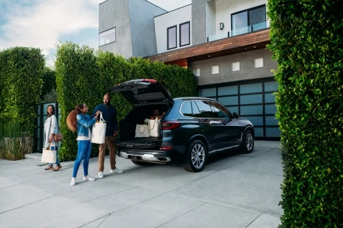 Family loads shopping bags into dark SUV parked outside modern house with green hedges and garage door.