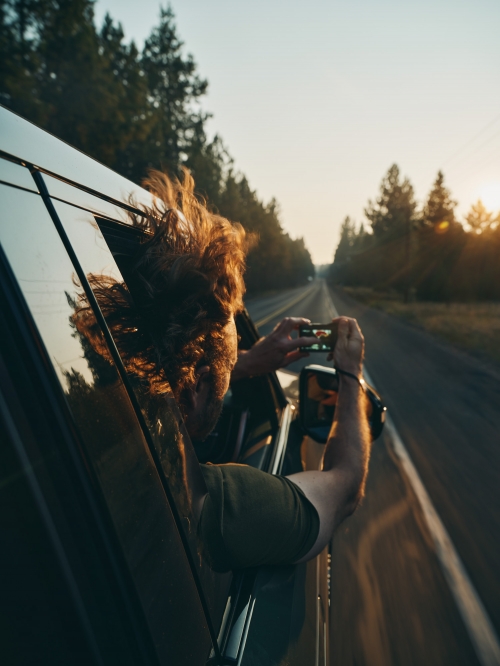 Person with curly hair leans out of a car window on a road trip, holding a smartphone to take a photo of the sunset and trees.