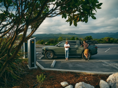 Two men pose with a Rivian electric truck charging at a station surrounded by greenery and a mountain landscape backdrop.