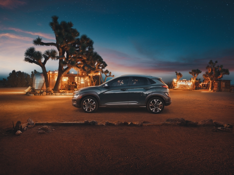 A gray Hyundai Kona is parked in a desert landscape at twilight, with Joshua trees and vintage trailers in the background under a starry sky.
