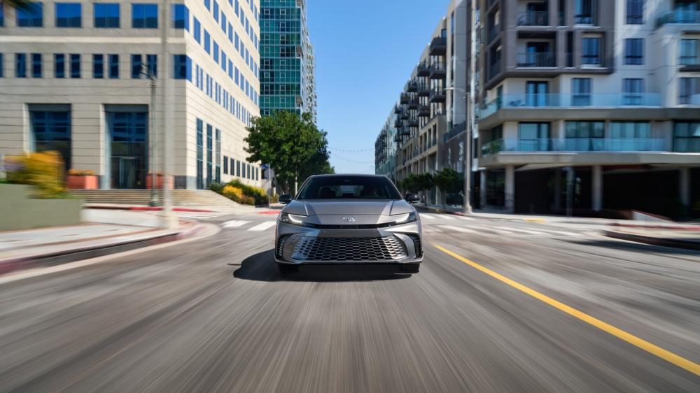 Silver car drives through a city street with modern buildings under a bright sky, showcasing motion blur and urban environment.