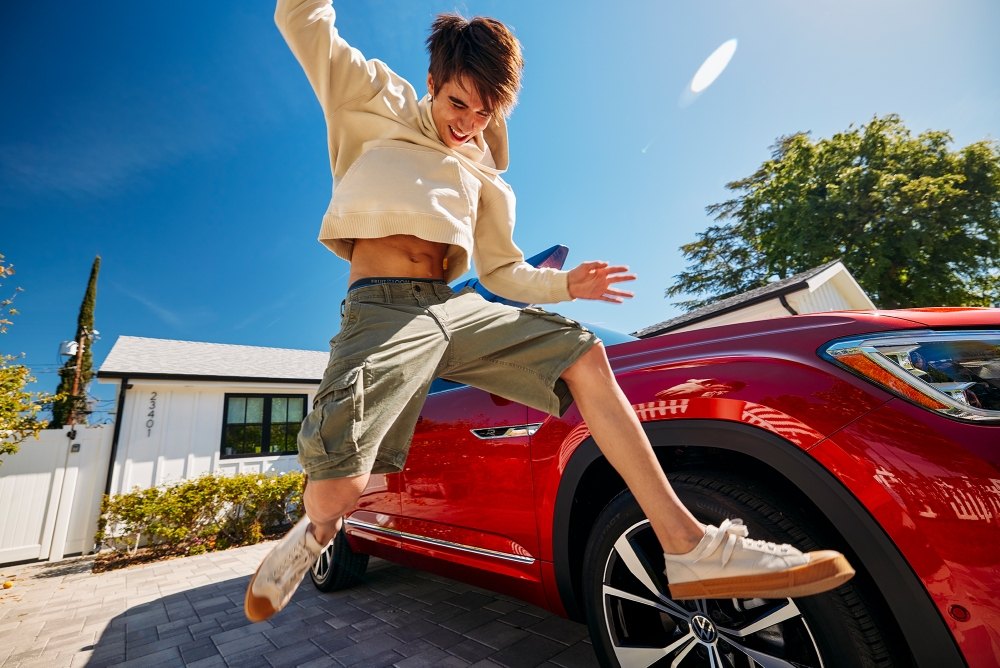Joyful young adult leaps high against a vibrant red car and clear blue sky, showcasing youthful energy and automotive style in an outdoor setting.