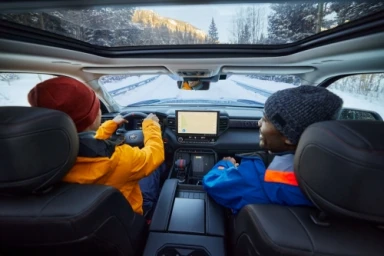 Two people in winter gear inside a car driving on a snowy mountain road viewed through a panoramic sunroof