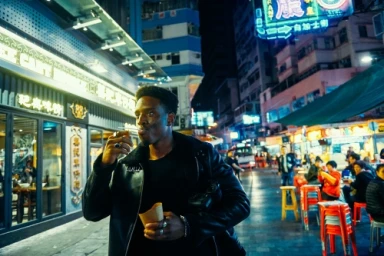 Black man in a black leather jacket eating street food at night on a bustling urban street lit by neon signs.