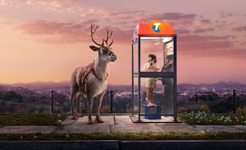 A child uses a phone booth next to a reindeer against a sunset city backdrop, blending fantasy with urban scenery in a surreal visual.