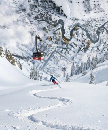 A skier carves down a snowy slope with a ski lift overhead and a snow-covered village nestled in the valley below on a sunny winter day.