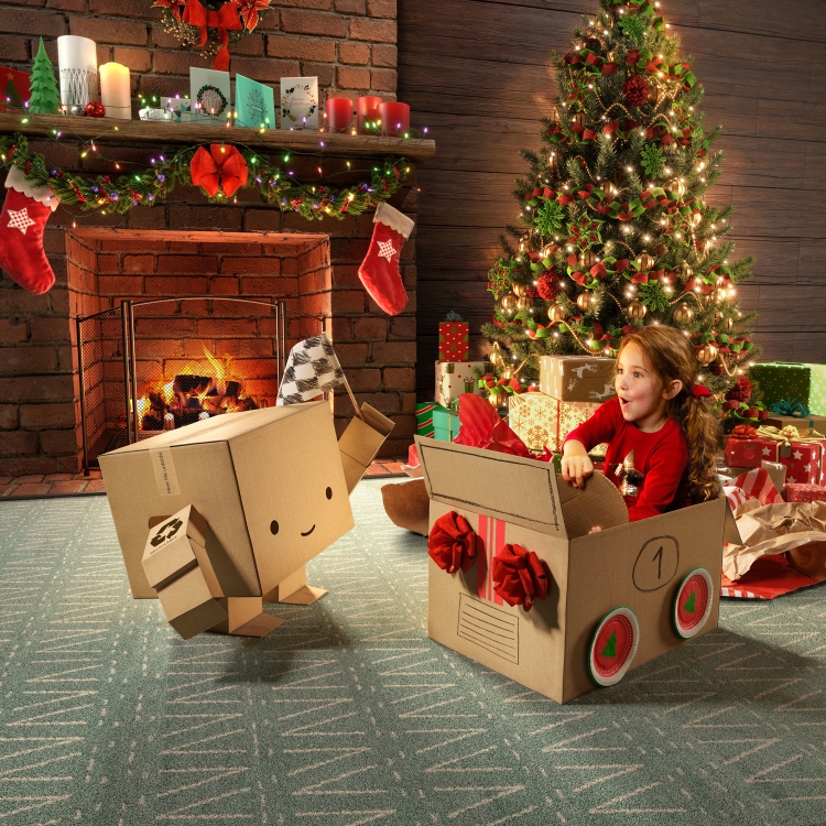 Young girl in a cardboard box 'car' interacts with a cardboard box character near a Christmas tree and decorated fireplace indoors.