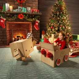 A young girl in a red sweater sits inside a cardboard box car next to a cardboard robot holding a checkered flag near a fireplace and Christmas tree.