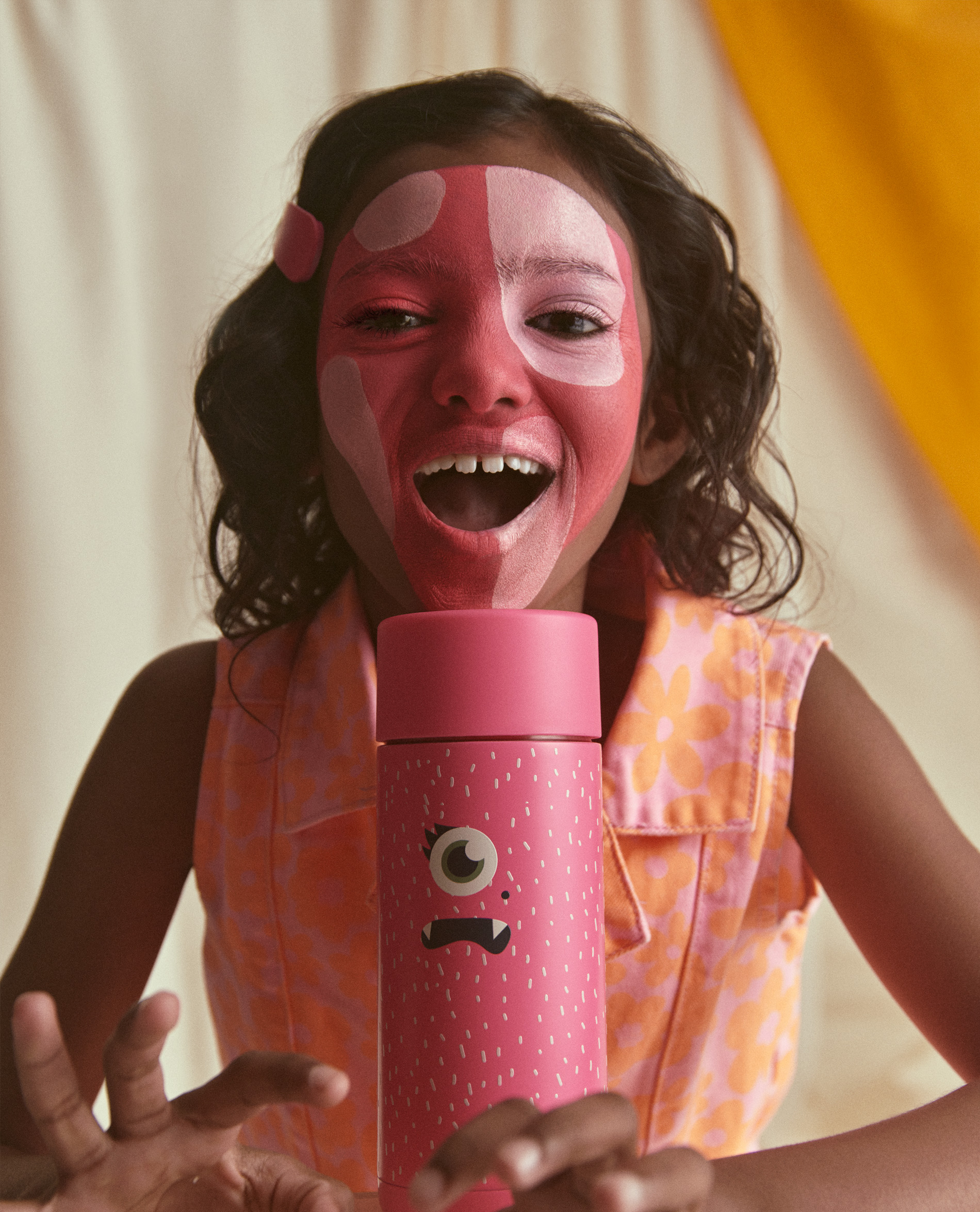 Young girl with painted face poses with pink bottle featuring cartoon face graphic in front of cream and yellow background.