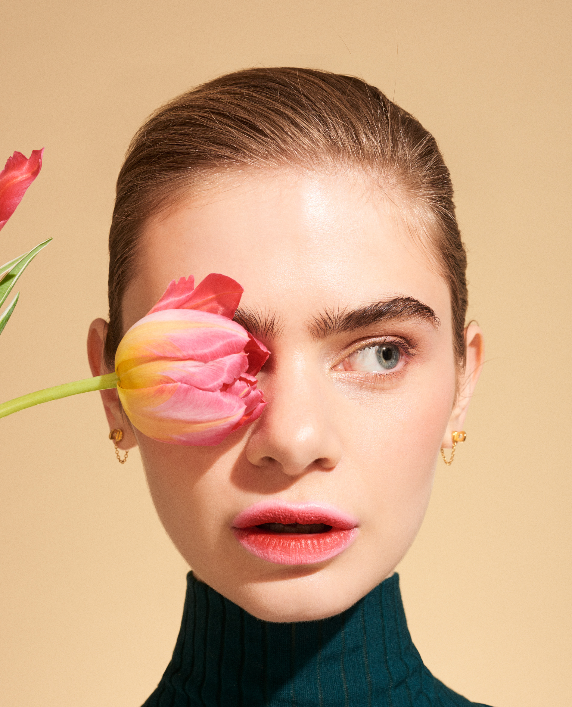 Close-up portrait of a fair-skinned young woman with a tulip flower covering one eye in a studio setting with a neutral backdrop.