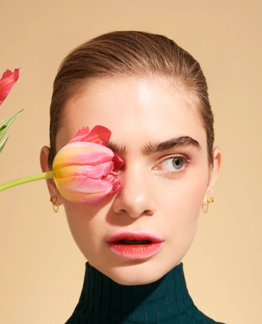 Close-up portrait of a fair-skinned young woman with a tulip flower covering one eye in a studio setting with a neutral backdrop.