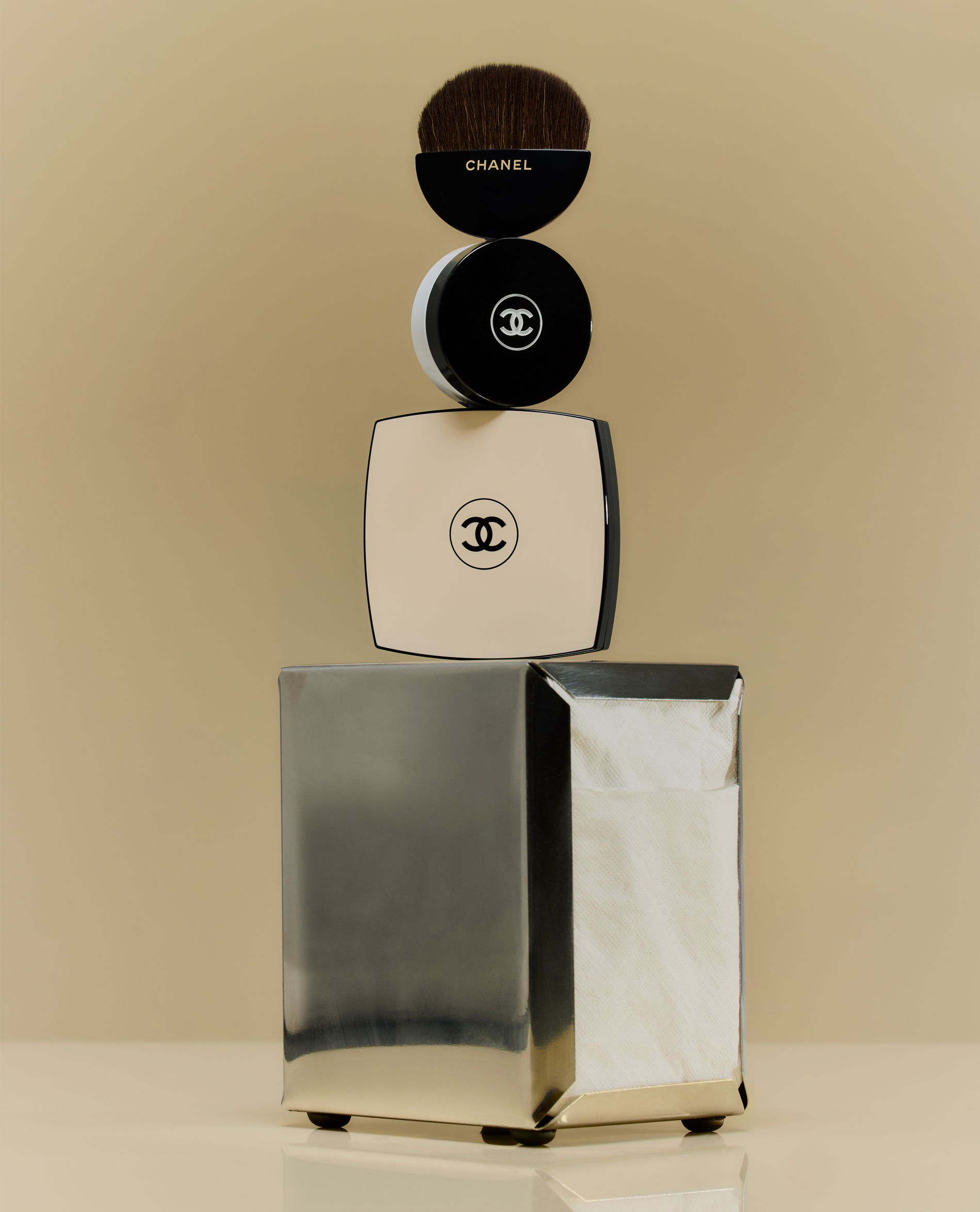 Close-up studio shot of Chanel makeup products stacked: a brush, round and square compacts on a wrapped silver box.