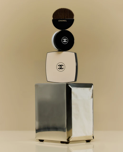 Close-up studio shot of Chanel makeup products stacked: a brush, round and square compacts on a wrapped silver box.