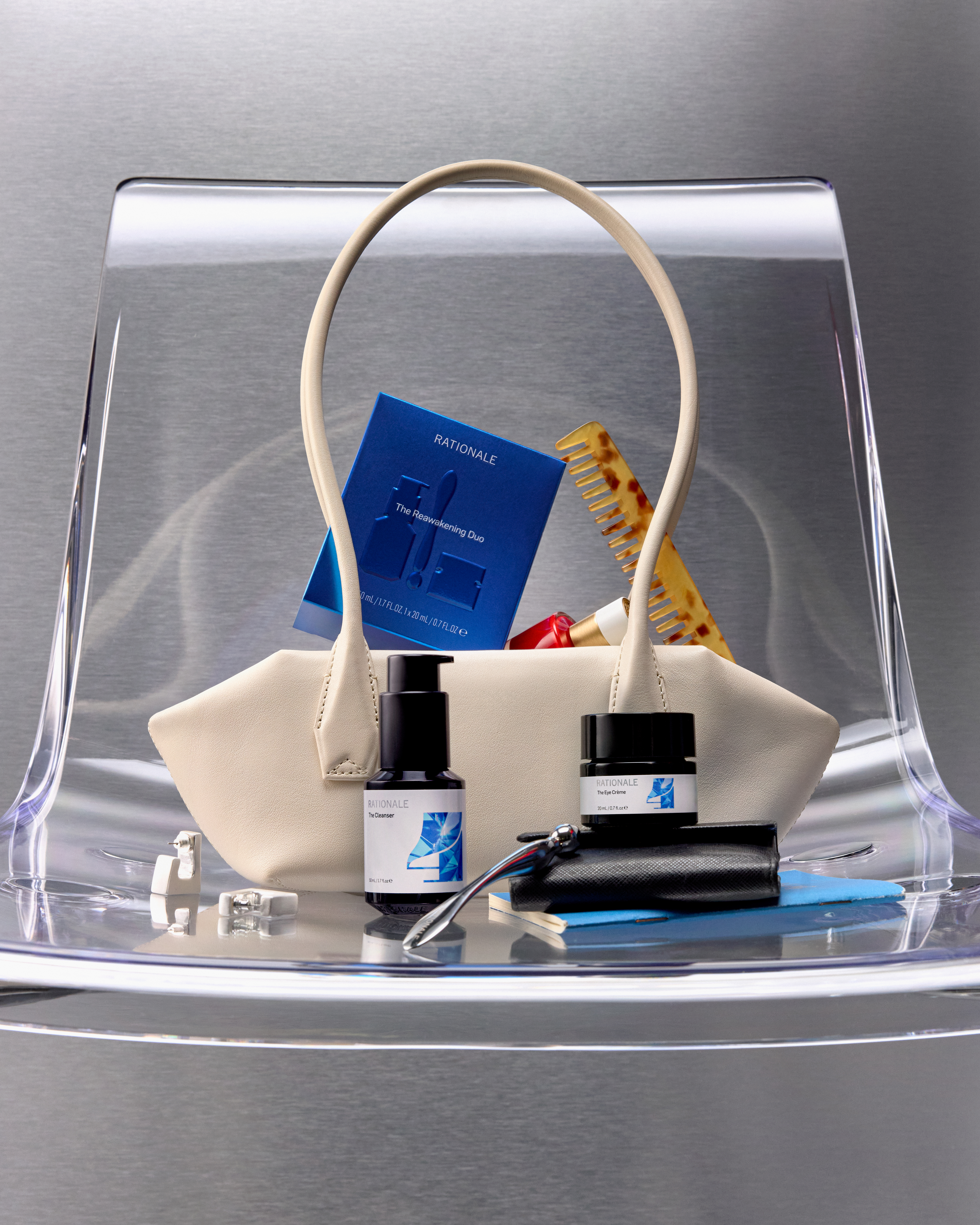 Fashion still life composition features a beige purse filled with beauty products, accessories, and notebook on a clear acrylic chair.