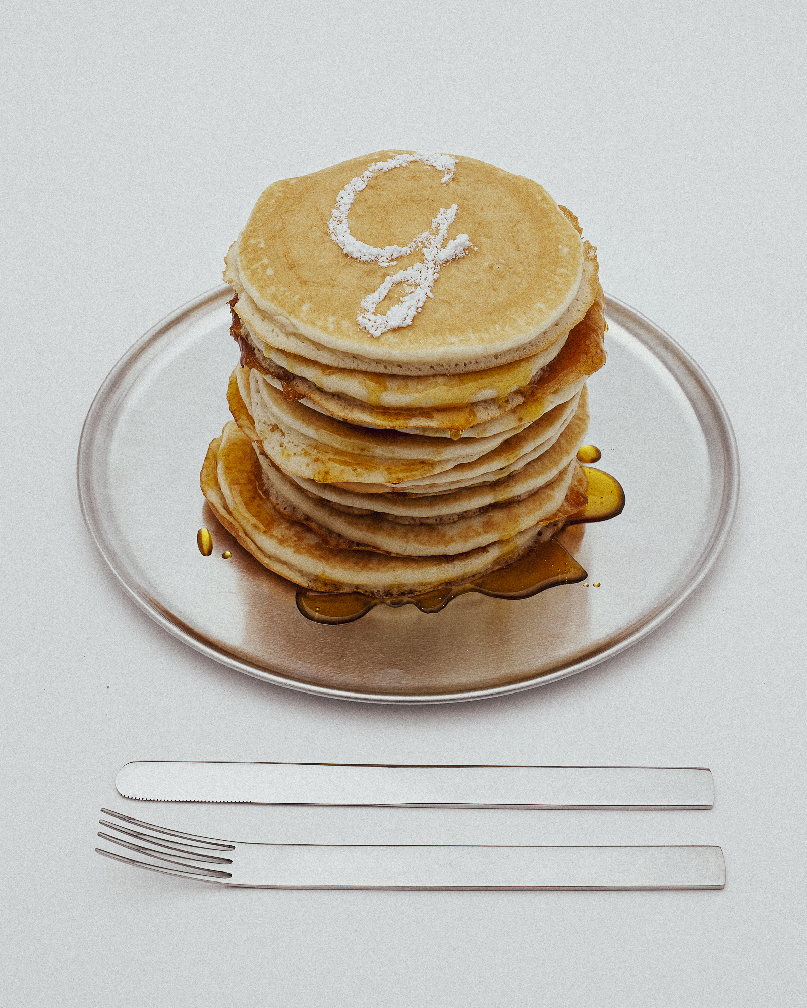 Stack of golden pancakes with syrup and powdered sugar 'G' on a silver platter, accompanied by a knife and fork on a white surface.