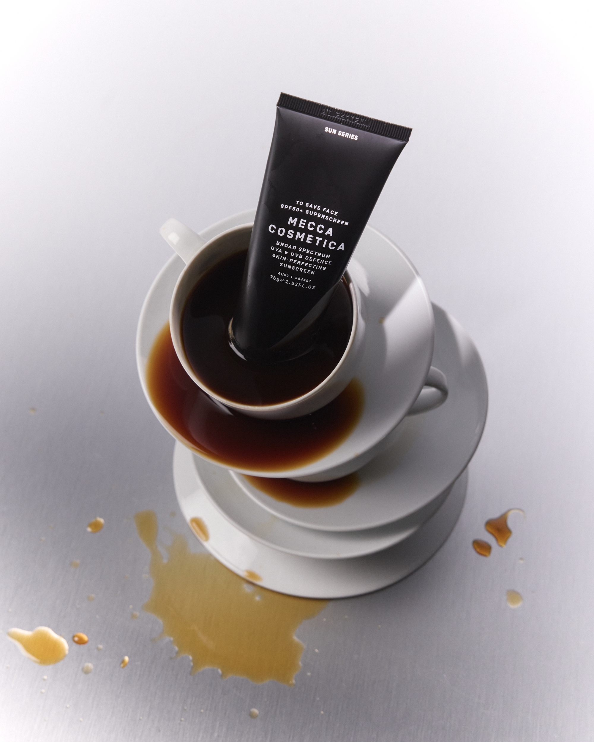 Mecca Cosmetica skincare tube in a coffee cup, which sits on a tower of white plates. Coffee is spilled on a silver table.