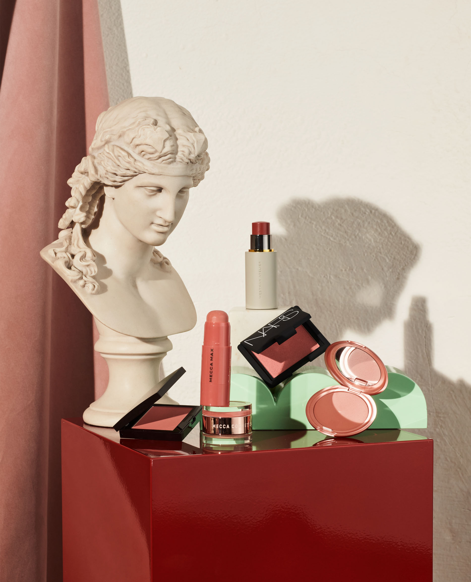 Still life composition featuring a marble bust, various cosmetic products like blushes and lipsticks, displayed on colorful geometric platforms.