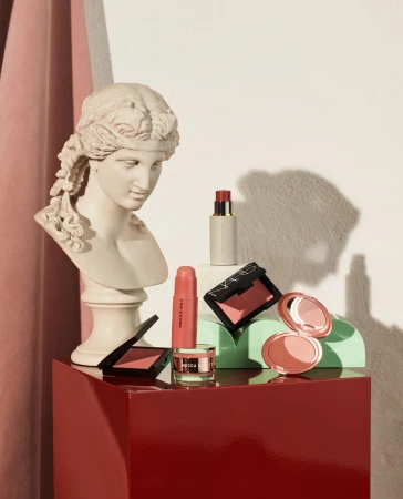 Still life composition featuring a marble bust, various cosmetic products like blushes and lipsticks, displayed on colorful geometric platforms.