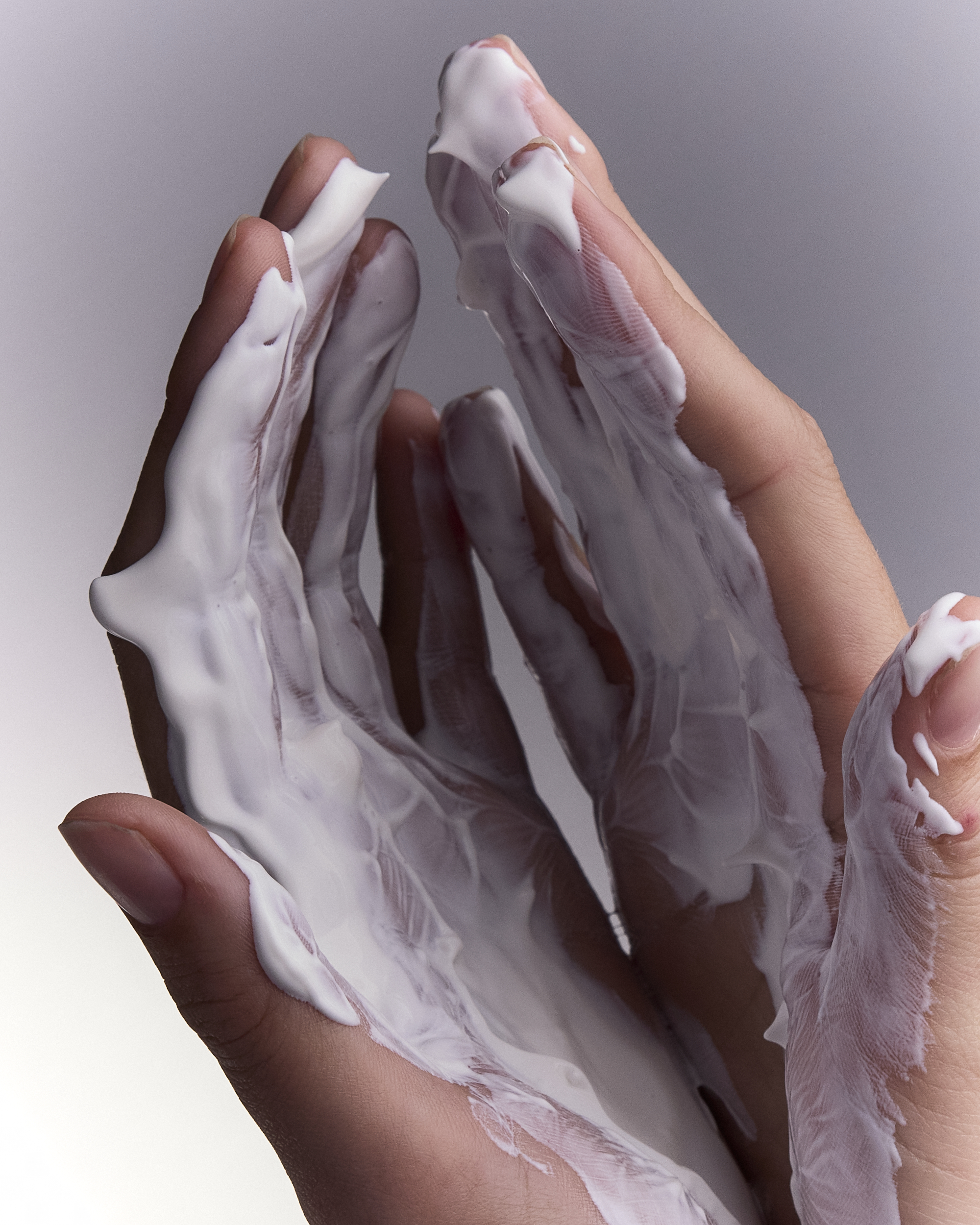 Close-up of two hands covered in white cream, showcasing skincare or beauty product application on a neutral background.