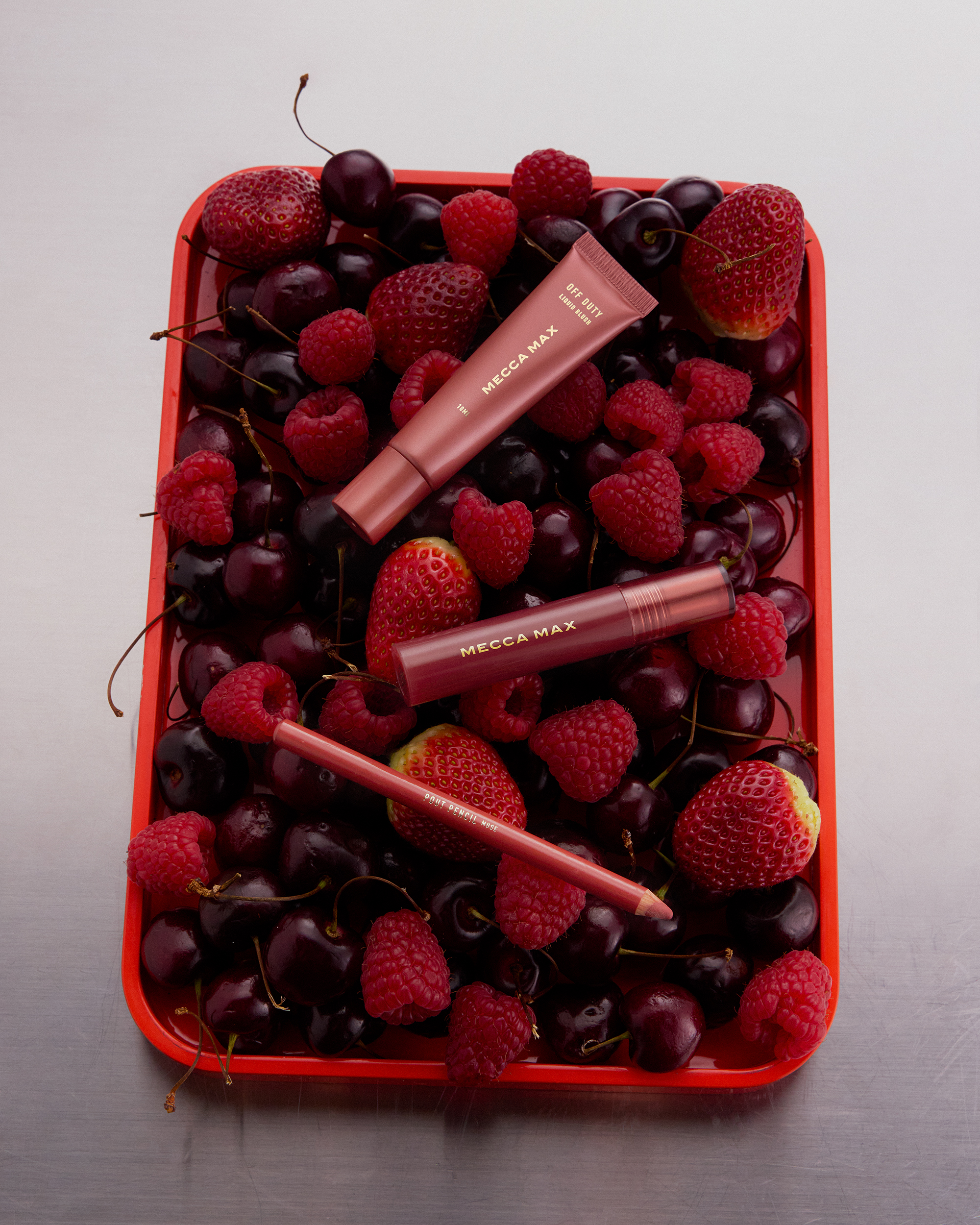 Still life featuring Mecca Max makeup products—eye tint, mascara, and lip pencil—arranged amidst fresh strawberries, raspberries, and cherries.