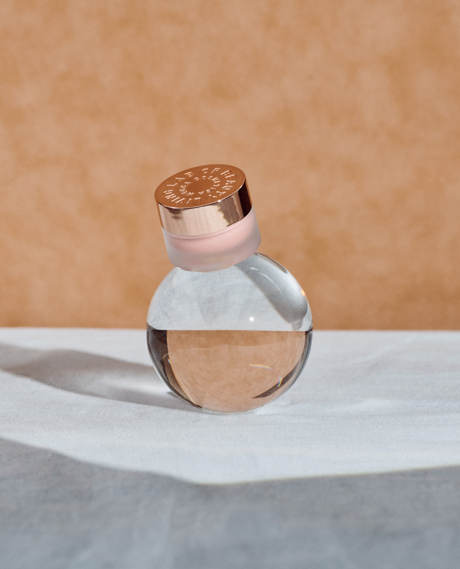 Close-up studio shot of a small, round glass perfume bottle with a metallic gold cap, set on a textured white surface against a tan background.