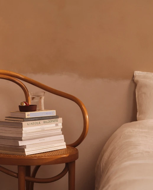 Stack of books on wicker chair next to bed. The books are topped with a small bowl and glass of water in a muted, neutral color palette.