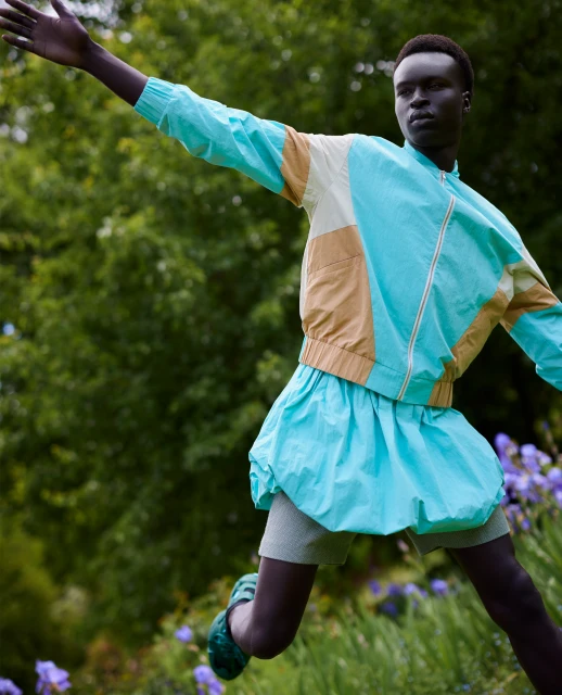 Fashionable Black model leaps in a turquoise bubble skirt, wearing a color-block jacket against a dreamy green backdrop with irises.