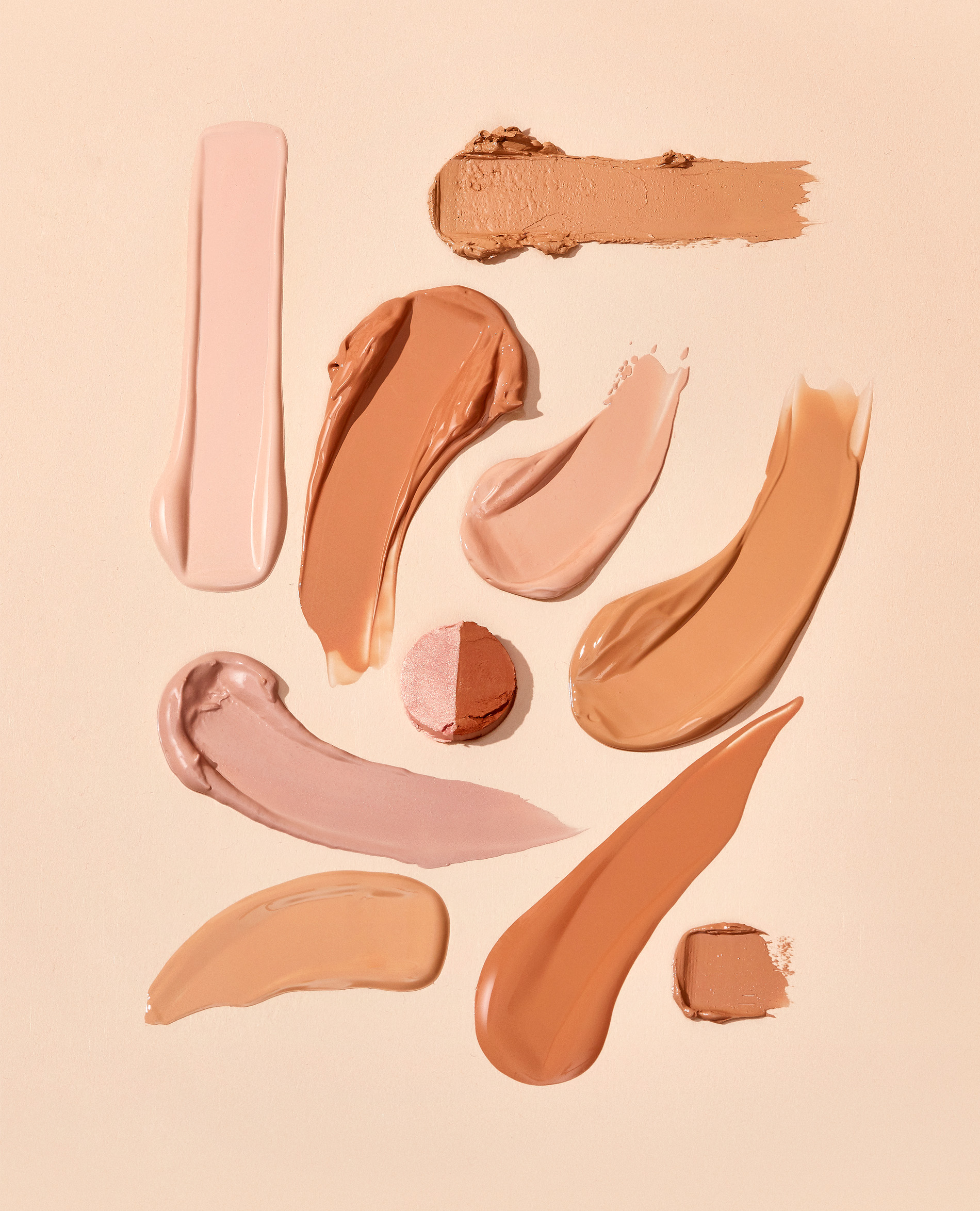 Cosmetics swatches in a variety of skin tone shades are displayed on a beige background, presented in a studio shot for beauty.