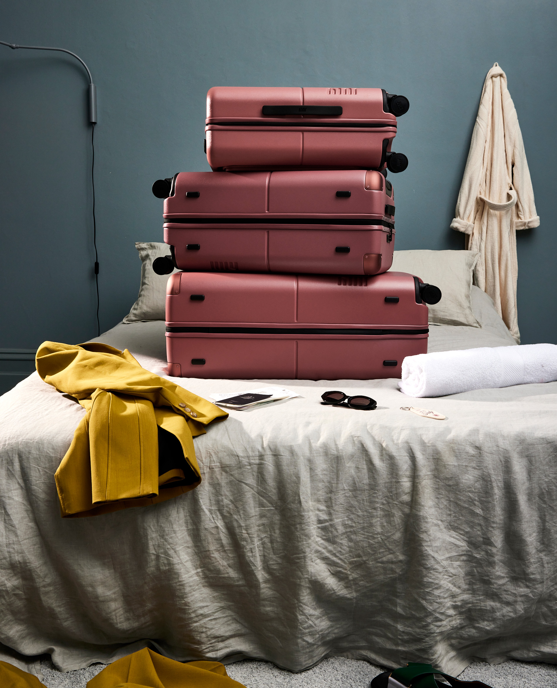 Four pink suitcases are stacked on a bed in a room with a grey-blue wall, a white bathrobe, a towel, and a yellow jacket.
