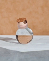 Cosmetic jar with a metallic rose gold cap balanced on a clear glass sphere over a white surface against a beige backdrop.