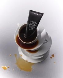 A black Mecca Cosmetica tube standing inside a white ceramic coffee cup with spilled dark liquid on stacked saucers and a metal table.