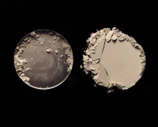 An empty circular cosmetic pan beside a cracked round beige pressed powder cake resting on a solid black background.