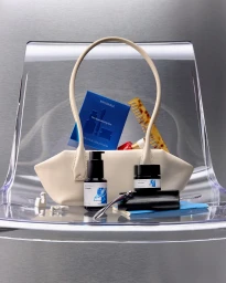 Beige leather handbag filled with beauty products resting on a clear acrylic chair against a textured grey background.